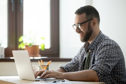 Happy male office worker smiling typing message at laptop, chatting with friends, writing positive email or consulting client online. Concept of laugher, positive work atmosphere, help and assistance